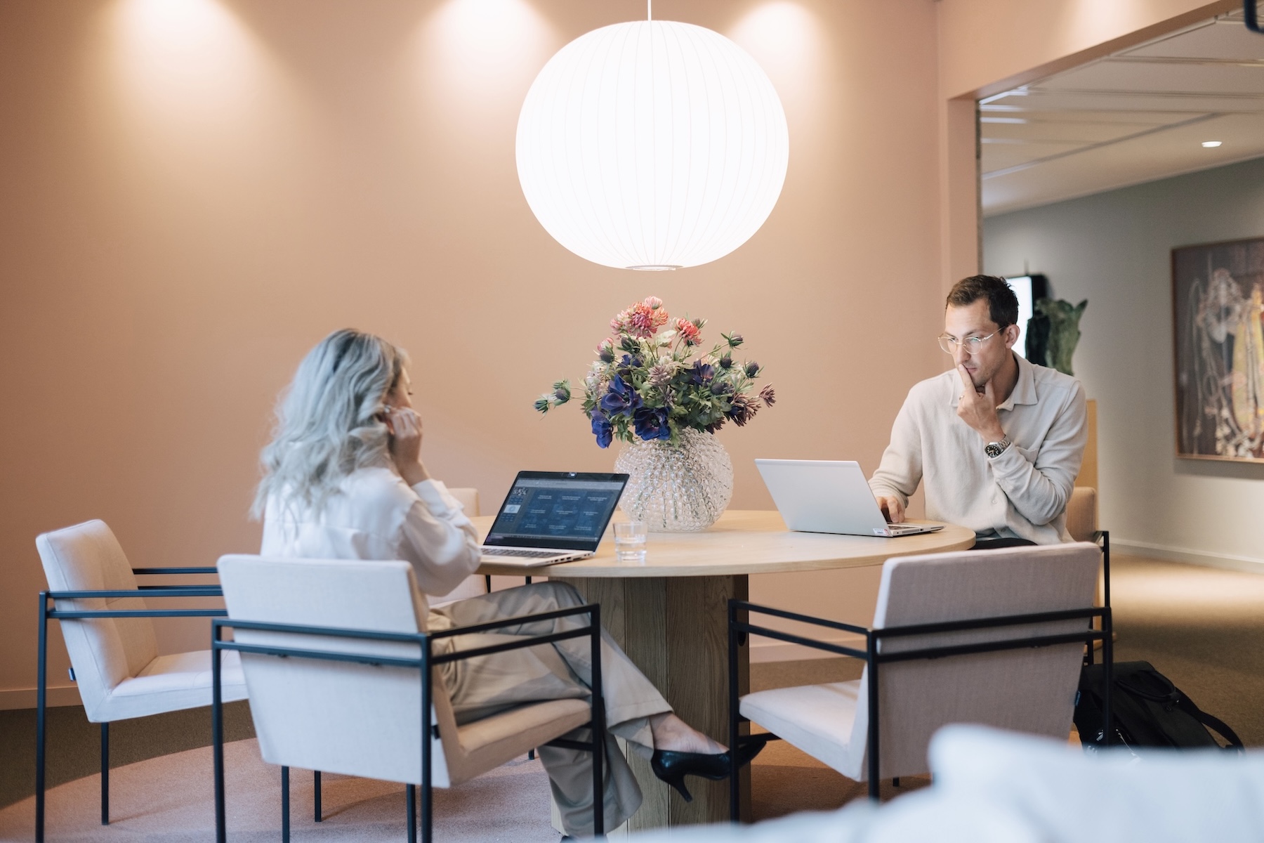 Två personer sitter vid ett runt bord med bärbara datorer på ett modernt kontor, en stor vit lampa över huvudet och en vas med blommor i mitten. Båda är fokuserade på sina skärmar, engagerade i arbete.
