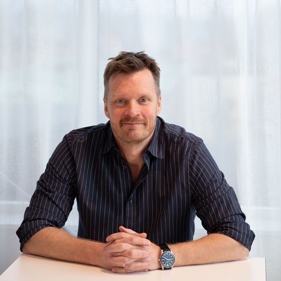 En man med kort hår och skägg sitter vid ett vitt bord med knäppta händer. Han bär en mörk, randig skjorta och ett armbandsur, med en vit gardin som bakgrund.