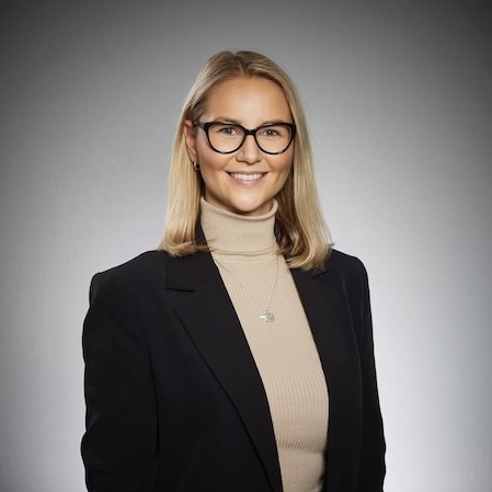 En kvinna med rakt blont hår och glasögon ler mot kameran. Hon är klädd i en beige polotröja och en svart kavaj och står mot en enfärgad grå bakgrund.