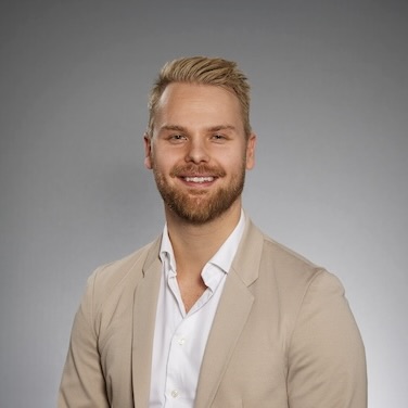 En man med kort blont hår och skägg ler mot kameran. Han är klädd i en ljusbeige kavaj över en vit skjorta och poserar mot en enfärgad, ljusgrå bakgrund.