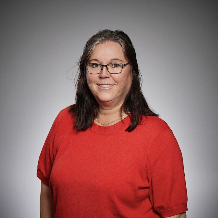 En kvinna med långt brunt hår och glasögon, klädd i en röd skjorta, ler mot kameran mot en grå bakgrund.