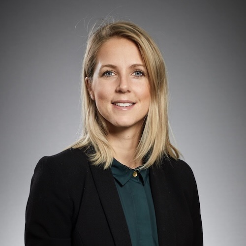 En kvinna med blont hår i svart kavaj och mörkgrön skjorta ler mot kameran mot en grå bakgrund.