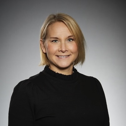 En kvinna med rakt blont hår, klädd i en svart topp, ler mot kameran mot en grå bakgrund.