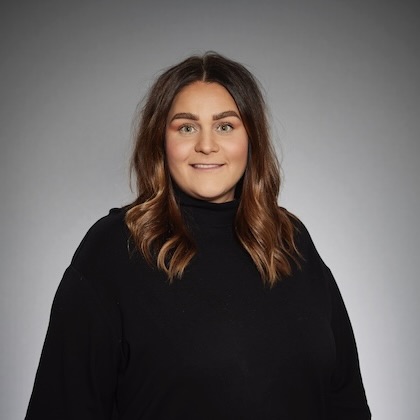 En kvinna med långt brunt hår och svart polotröja poserar framför en grå bakgrund och ler mjukt mot kameran.