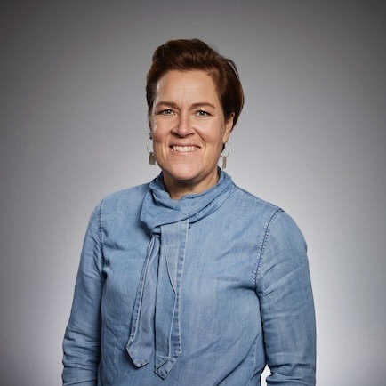 En kvinna med kort brunt hår, klädd i en ljusblå, långärmad blus med knytband i nacken, står och ler framför en grå bakgrund.