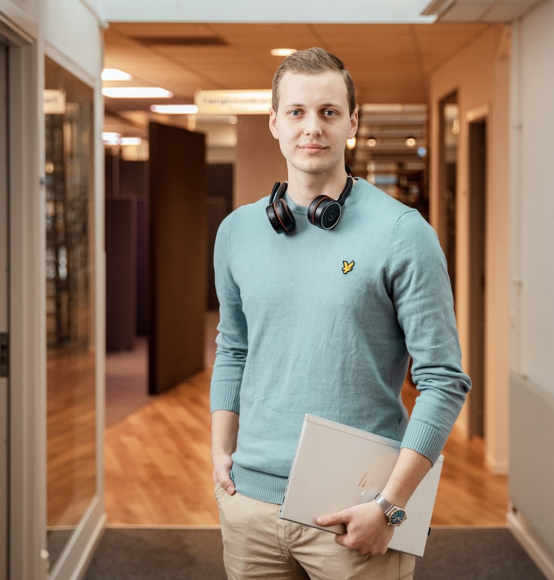 En ung man med kort hår, klädd i ljusblå tröja, khakifärgade byxor och hörlurar runt halsen står i en korridor med en stängd laptop och ler mot kameran.