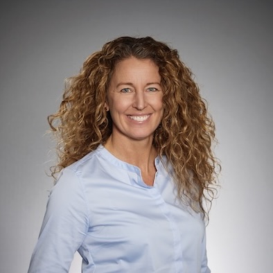 En kvinna med långt lockigt hår ler mot kameran. Hon är klädd i en ljusblå skjorta med knappar och poserar framför en enfärgad grå bakgrund.