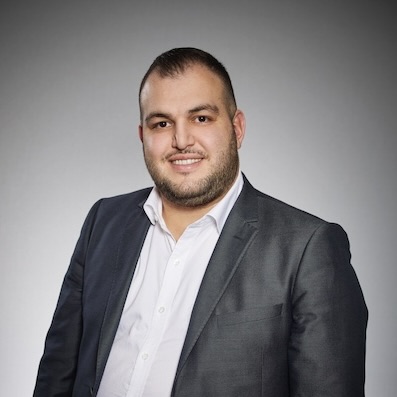 En man med kort mörkt hår och skägg, klädd i en mörk kostymjacka över en vit skjorta, ler mot kameran mot en neutral grå bakgrund.