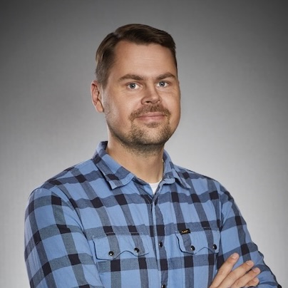 En man med kort brunt hår och ansiktsbehåring bär en blå och svart rutig skjorta, står med armarna i kors och ler lätt mot en grå bakgrund.