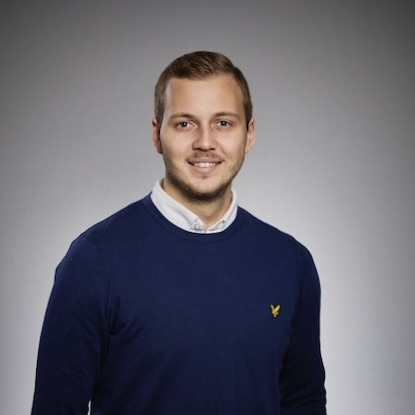 En ung man med kort ljusbrunt hår, klädd i en marinblå tröja över en vit skjorta med krage, ler mot kameran mot en grå bakgrund.