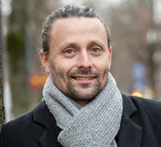 En man med ljusa ögon och bakåtkammat hår ler utomhus. Han bär en grå stickad halsduk och en mörk kappa, med en suddig bakgrund av träd och ljus.