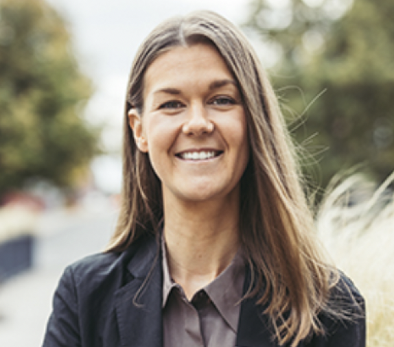 En kvinna med långt brunt hår, klädd i mörk kavaj och grå skjorta, ler utomhus med suddiga träd och växter i bakgrunden.