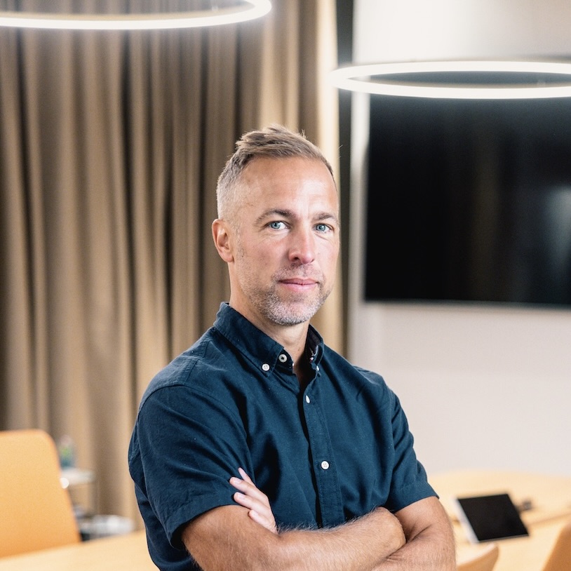 En man med kort ljust hår och vältrimmat skägg står med armarna i kors på ett modernt kontor. Han bär en kortärmad mörk skjorta. Bakom honom finns beige gardiner, en svart skärm och runda taklampor.