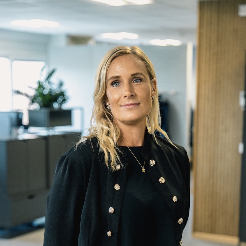 En kvinna med blont hår och svart kavaj står på ett modernt kontor och tittar in i kameran. I bakgrunden syns stora fönster, växter och kontorsmöbler i ett ljust, öppet utrymme.