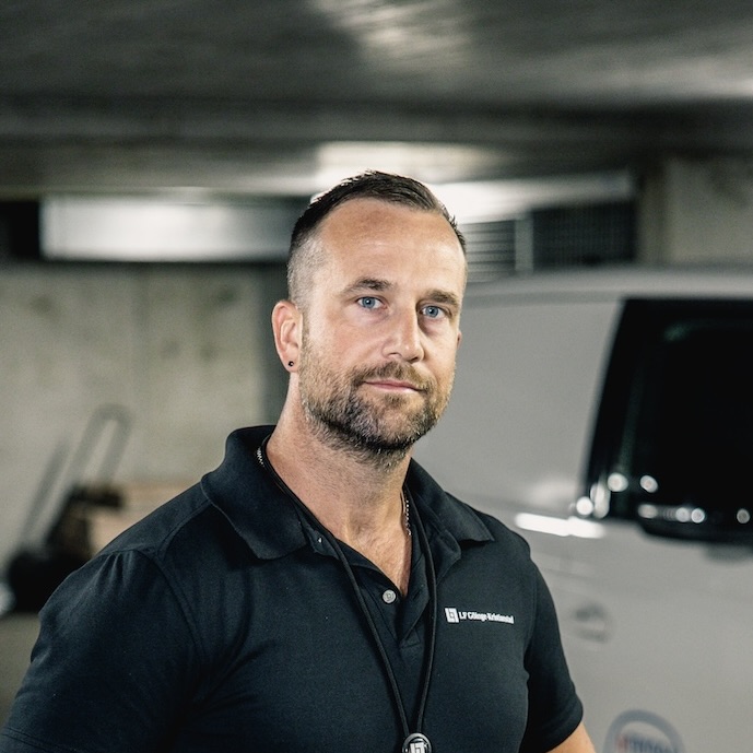 En man med kort hår, skägg och örhänge står i ett svagt upplyst parkeringsgarage, klädd i svart skjorta med krage och nyckelband. I bakgrunden syns ett vitt fordon och betongväggar.