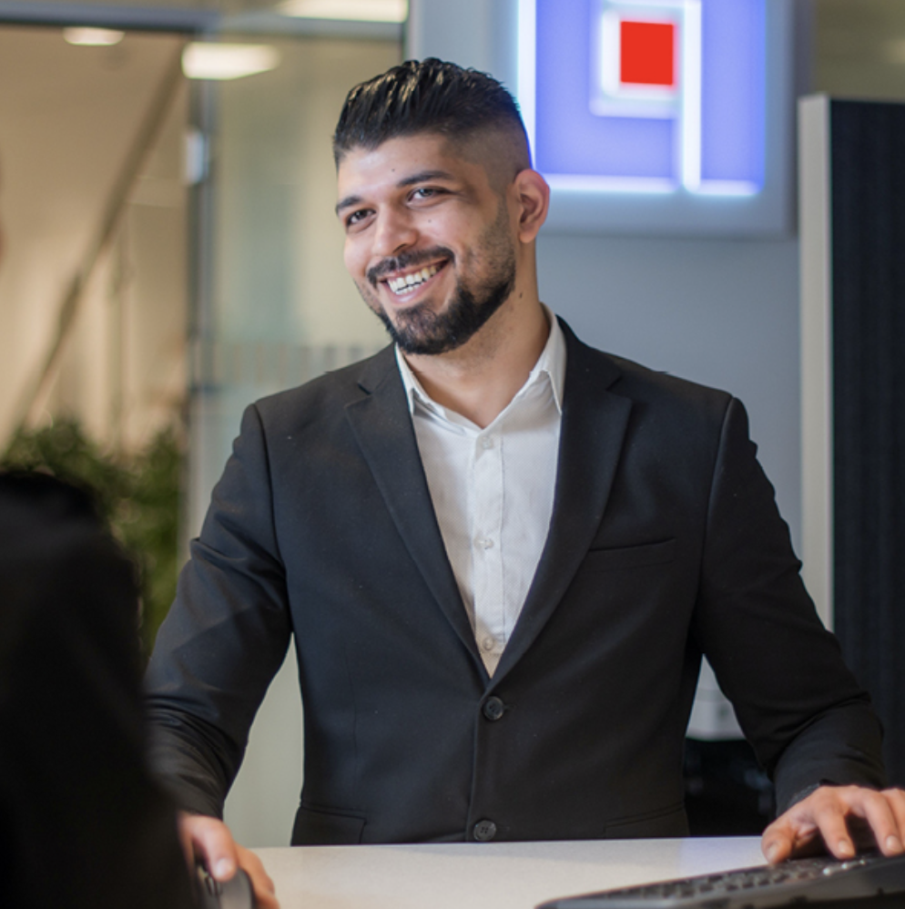 En man med kort skägg och bakåtslickat hår, klädd i svart kostym och vit skjorta, ler när han står vid en receptionsdisk i en modern kontorsmiljö.