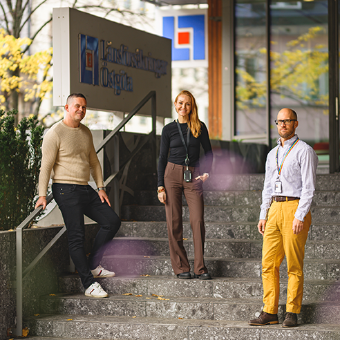 Tre personer står på en stentrappa utomhus nära en entré till en byggnad med en skylt med texten "Länsförsäkringar Östgöta". Träd med gula blad syns i bakgrunden. Alla tre ler och är vända mot kameran.