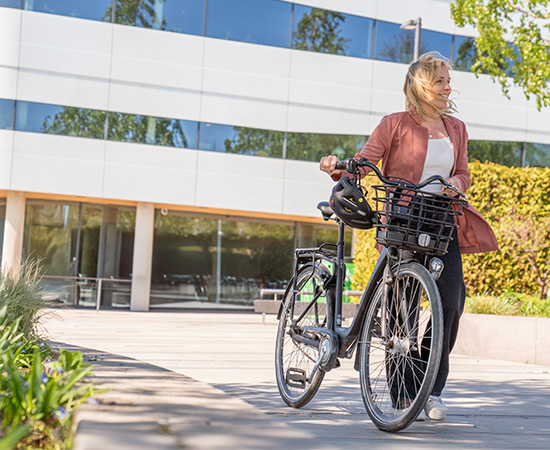 En kvinna med blont hår går bredvid en cykel en solig dag framför en modern glasbyggnad, håller i styret och tittar åt sidan.