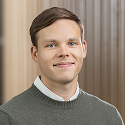 En kvinna med rakt, axellångt mörkt hår och ljus hy, klädd i en marinblå topp, står framför en ljusbrun, vertikalt randig bakgrund och tittar in i kameran med ett neutralt uttryck.