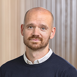 En man med kort brunt hår och vältrimmat skägg, klädd i en rödvitrutig skjorta, ler mot kameran mot en suddig inomhusbakgrund.